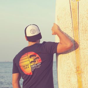 Ragazzo con tavola da surf sulla spiaggia, che indossa la t-shirt blu navy con stampa sulla schiena Surf Rider 1984 Costa Est